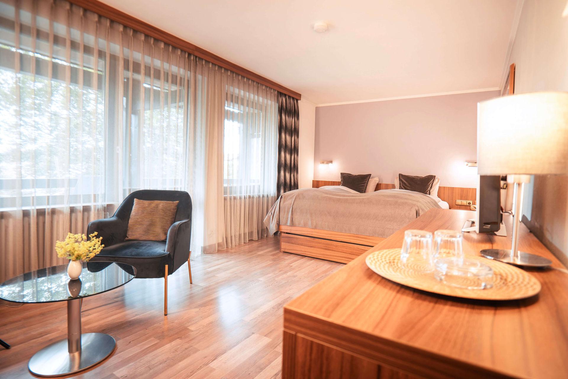Helles Hotelzimmer mit großem Bett, Sessel, Holztisch und Fenster mit Vorhängen.