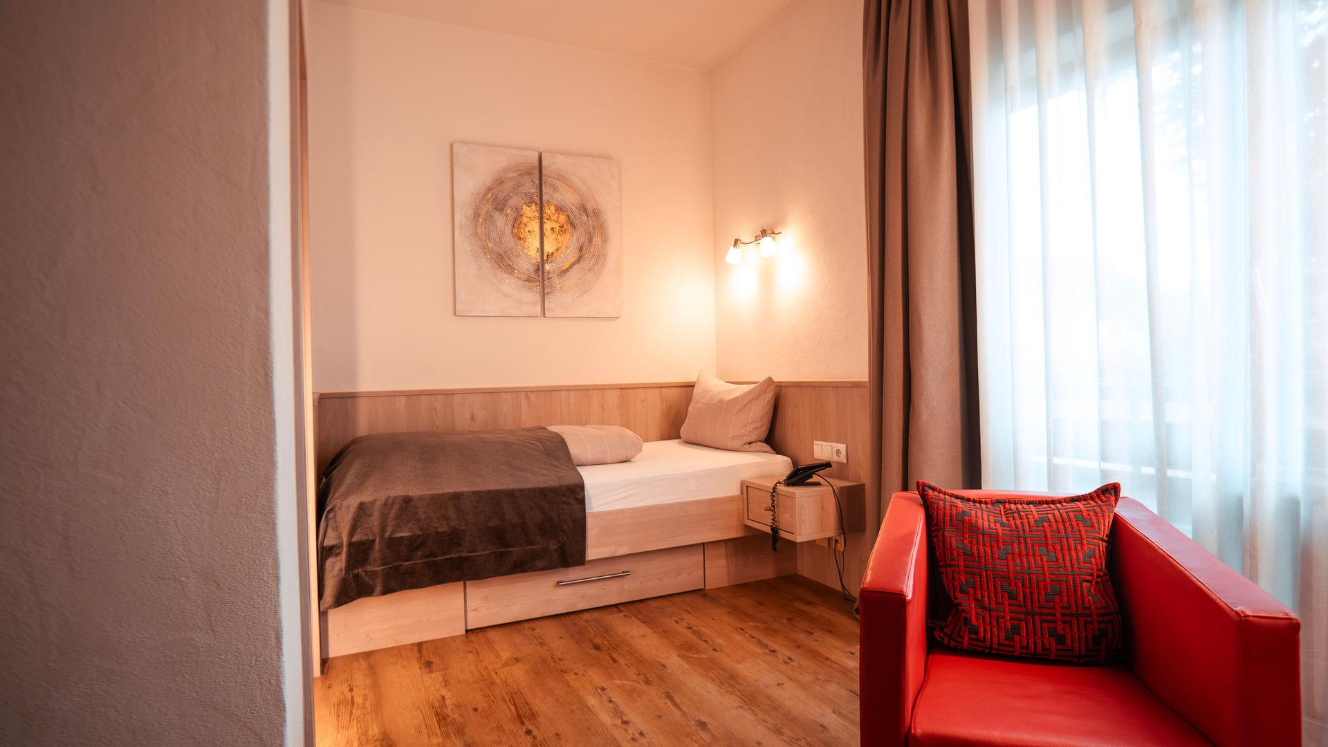 Cosy single room with a narrow bed, wooden floor, red armchair and curtains.
