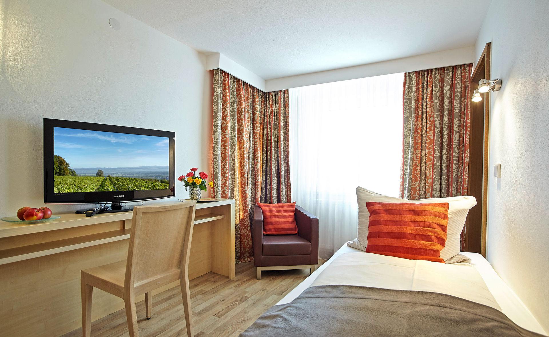 Bright single room with a single bed, desk, television and armchair.