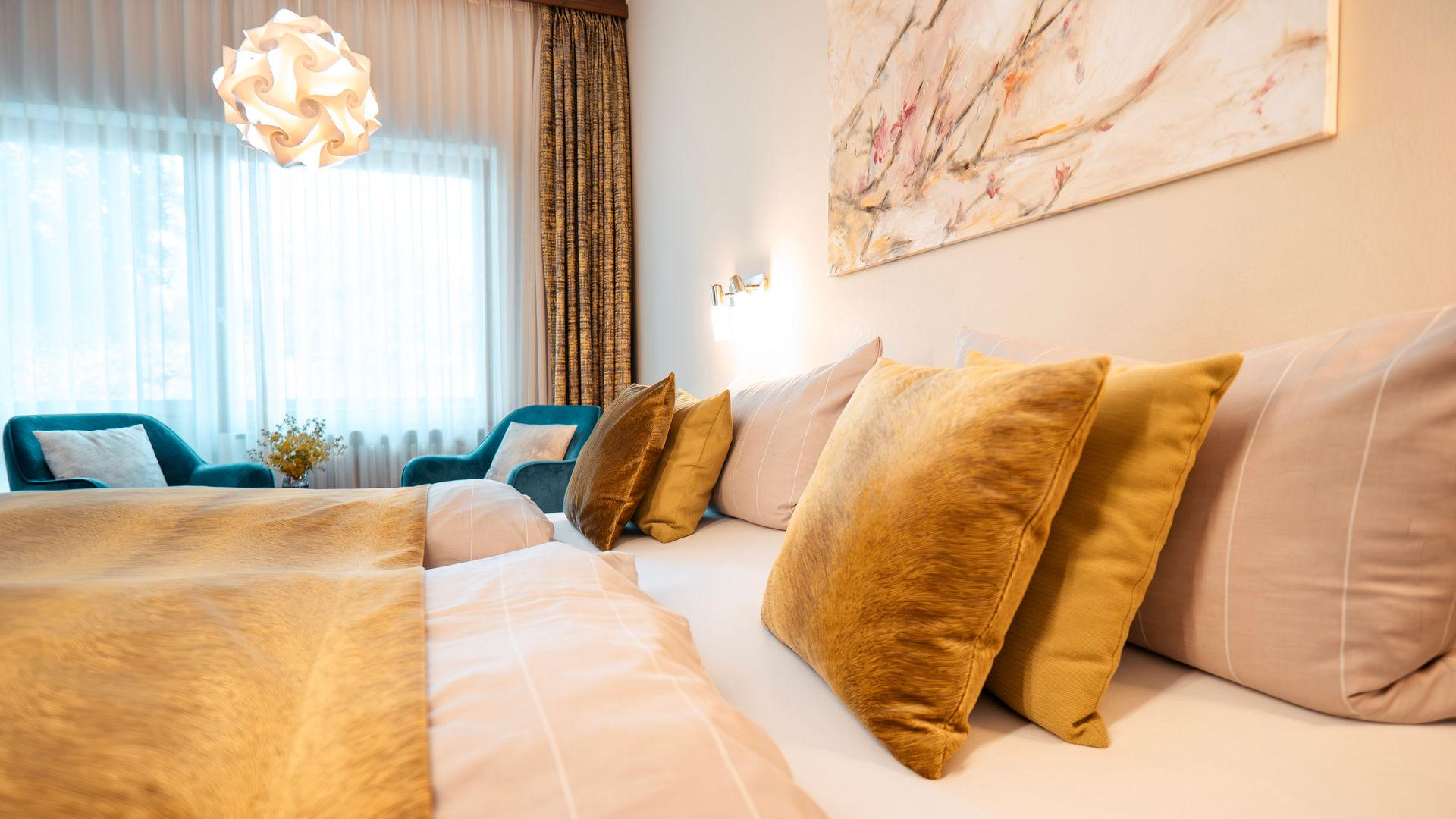 Gemütliches Hotelzimmer mit großem Bett, goldenen Kissen und Sesseln am Fenster.