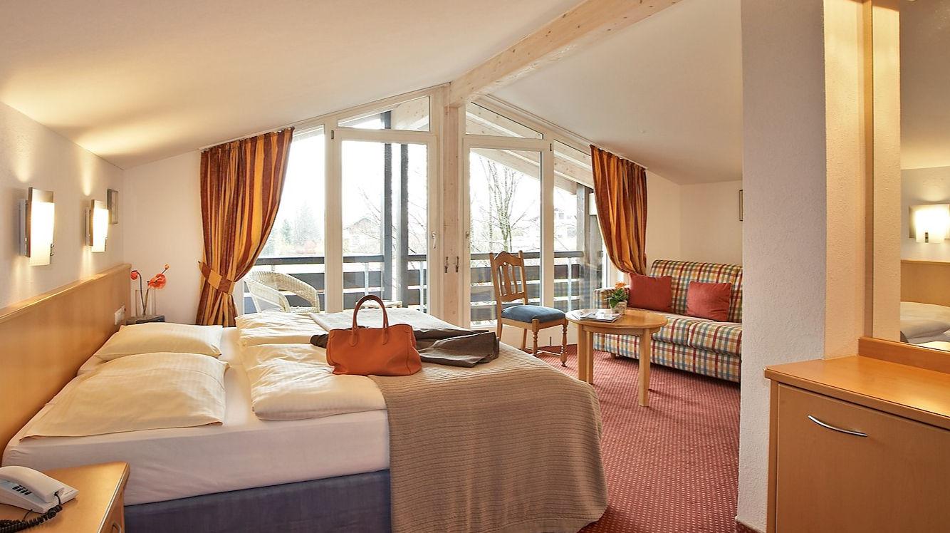 Helles Hotelzimmer mit großem Bett, Sofa, Balkonzugang und rotem Teppich.