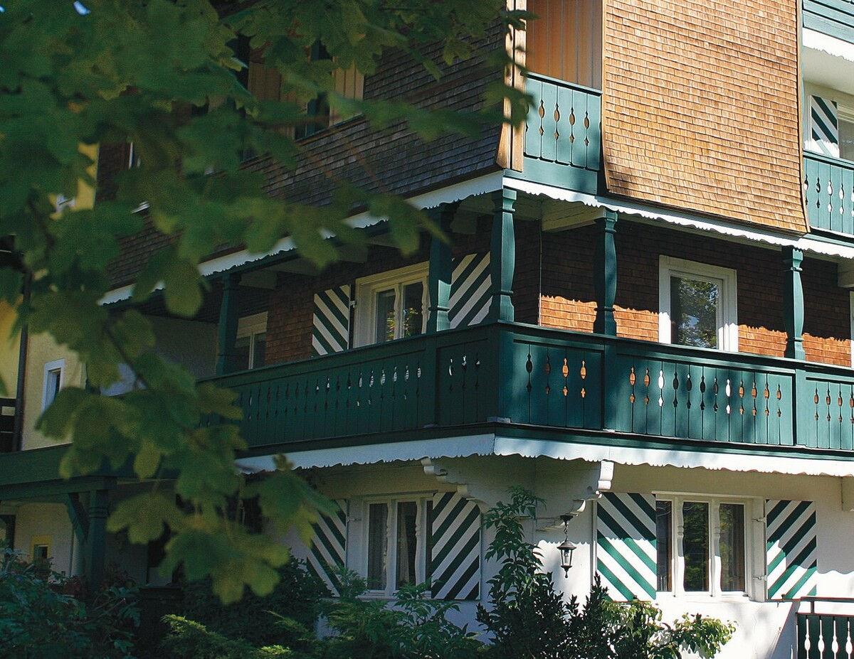 Hotel façade with a green wooden balcony framed by foliage.