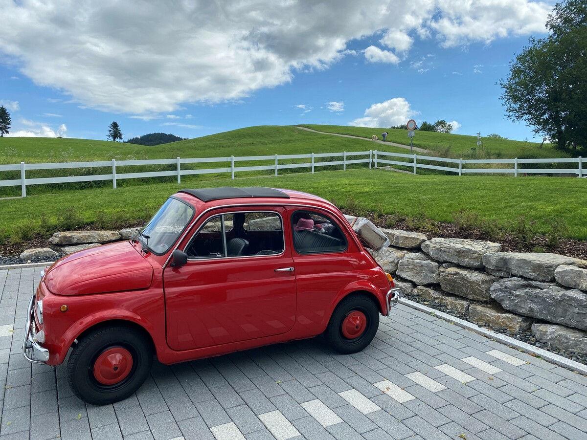 Red vintage Fiat 500 on the paved hotel driveway in front of green hills.