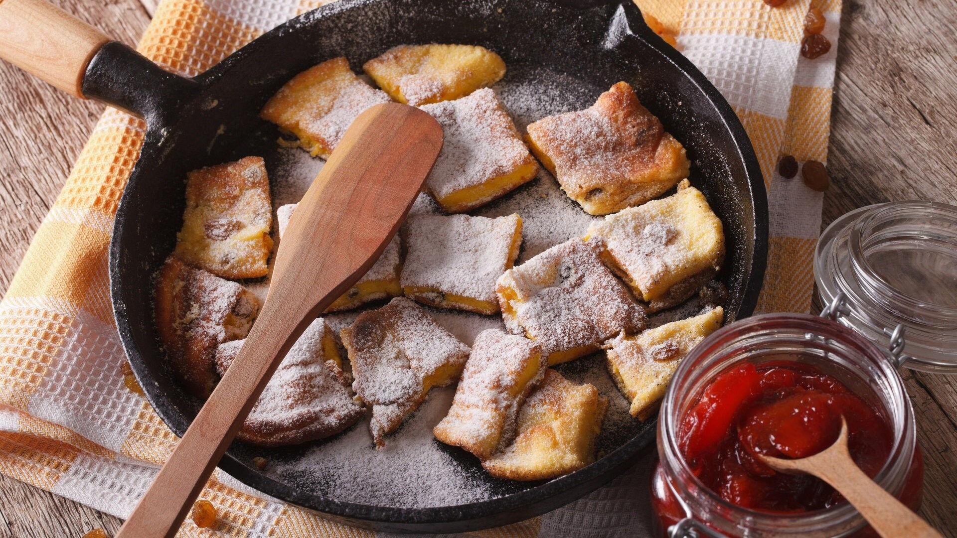 Goldbrauner Kaiserschmarrn mit Puderzucker in Pfanne, dazu rote Marmelade.