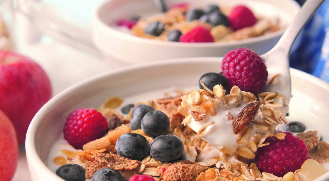 Löffel nimmt Müsli mit Joghurt, Blaubeeren und Himbeeren aus weißer Schüssel.
