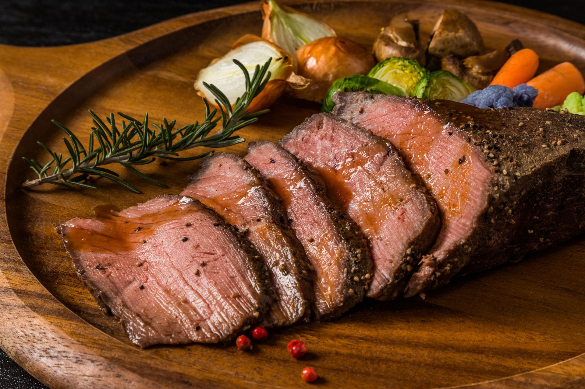 Saftiger, aufgeschnittener Rinderbraten mit Rosmarin und geröstetem Gemüse auf Holzplatte.