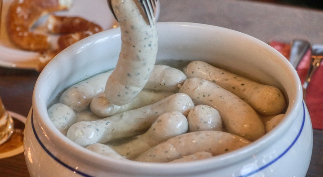Eine Weißwurst wird mit Zange aus warmem Topf gehoben, bayerische KULINARIK.
