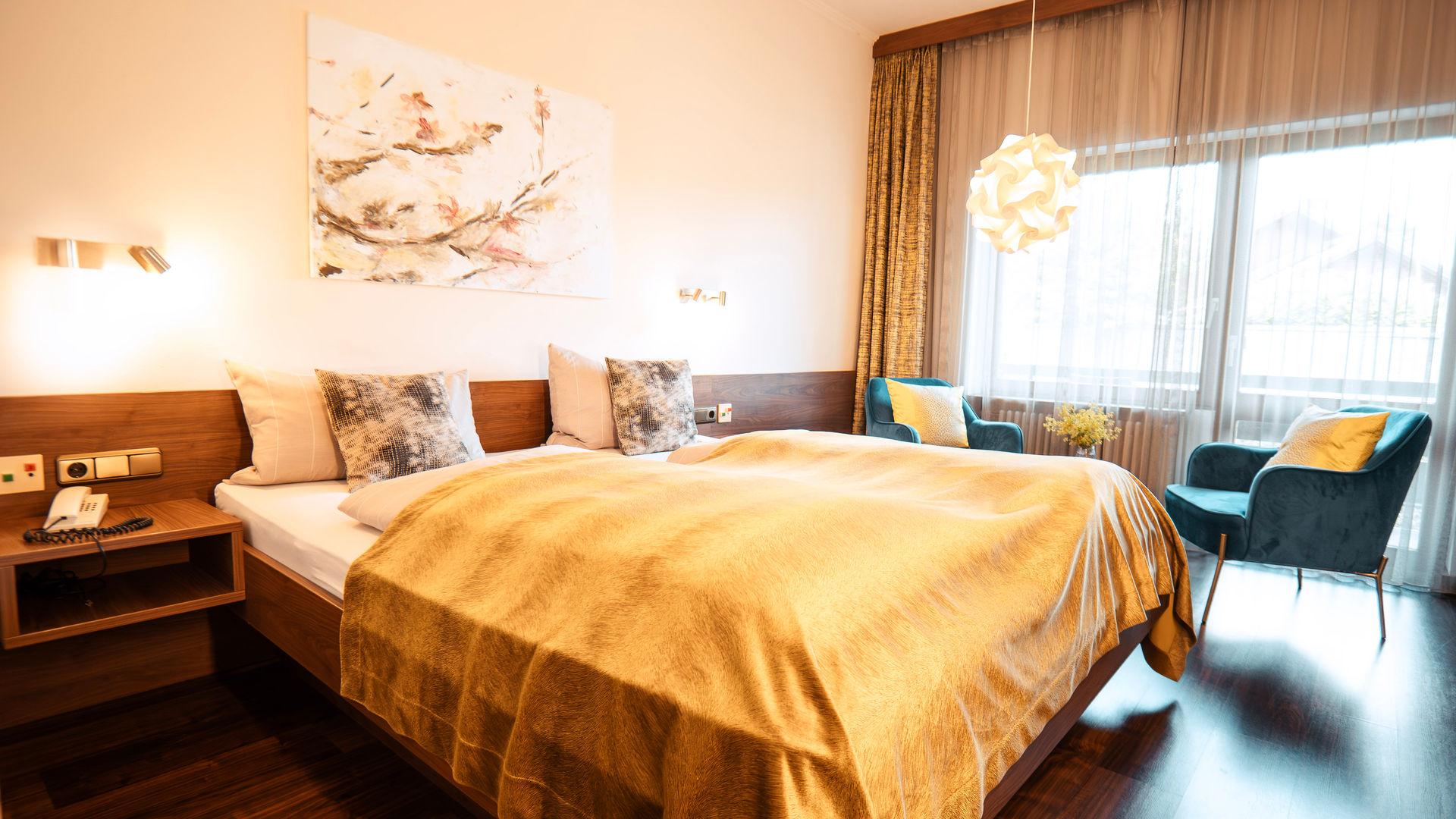 Double room with a golden bedspread, blue armchairs by the window and a spherical pendant lamp.